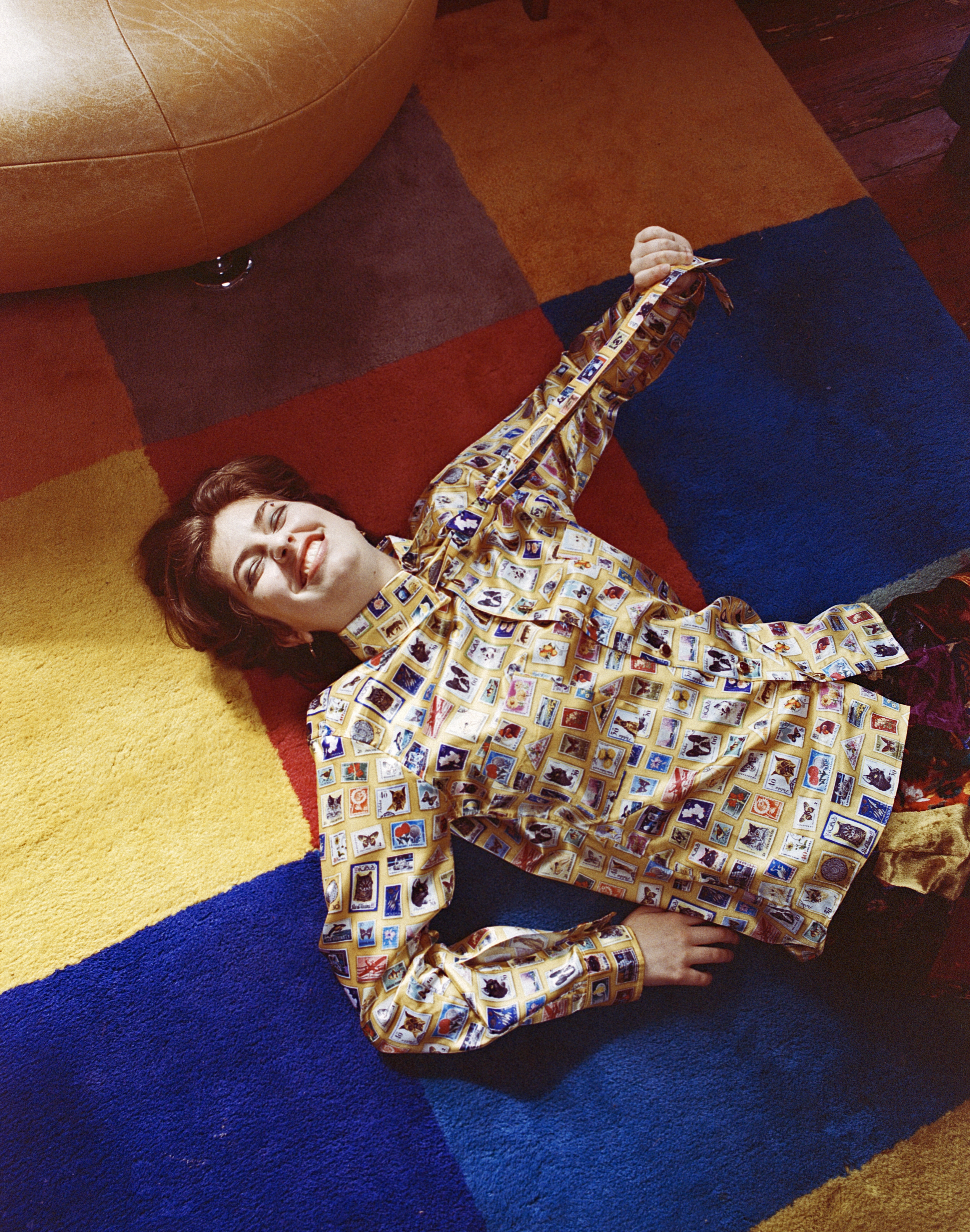 A white androgynous person in a yellow patterned shirt and tie lays on a rug with colorful rainbow squares